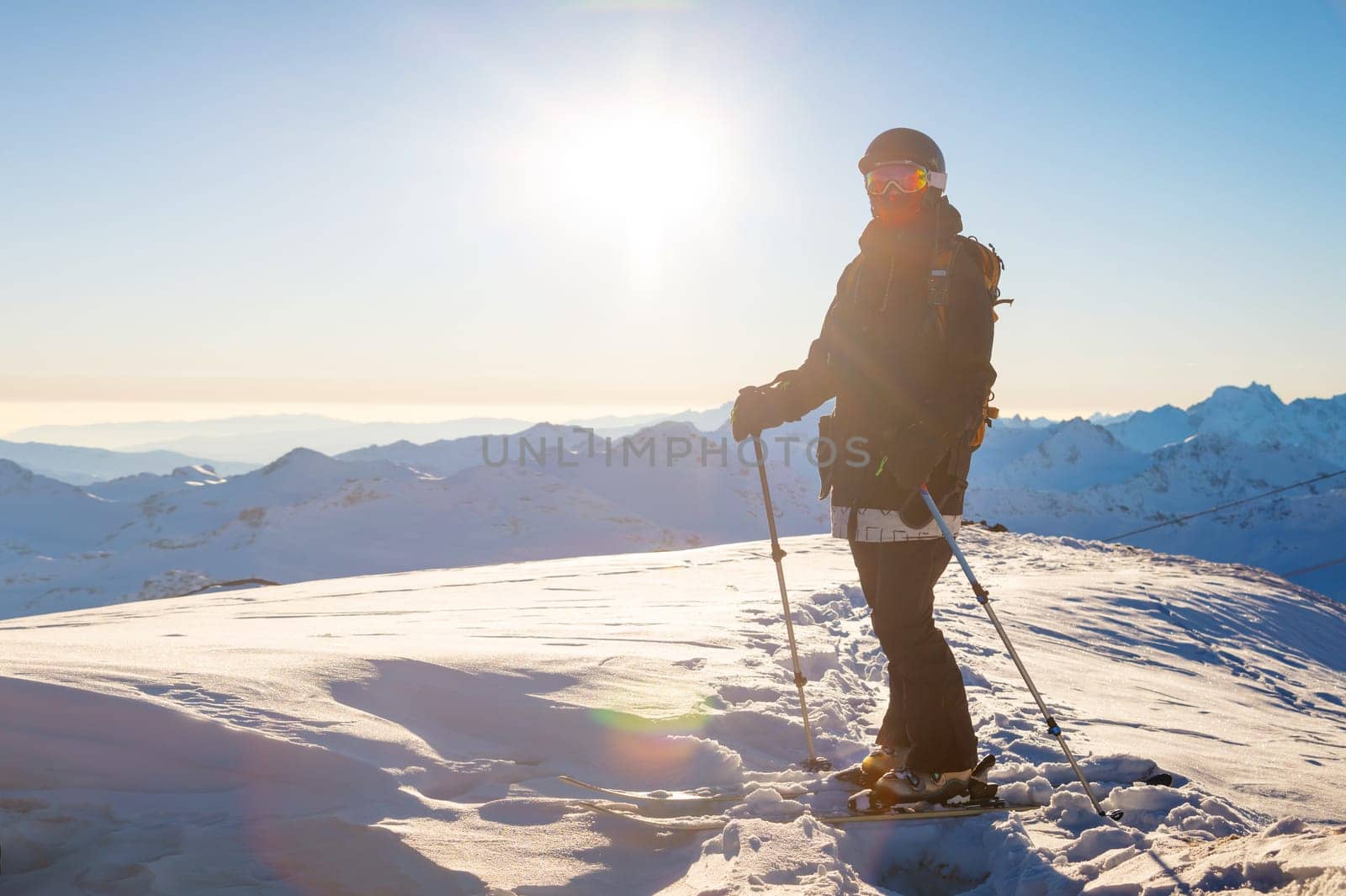 An adult girl on skis stands on a mountainside, admiring the mountain landscape, looking at the camera and enjoying the sunny winter weather. Interesting winter activity. side view.