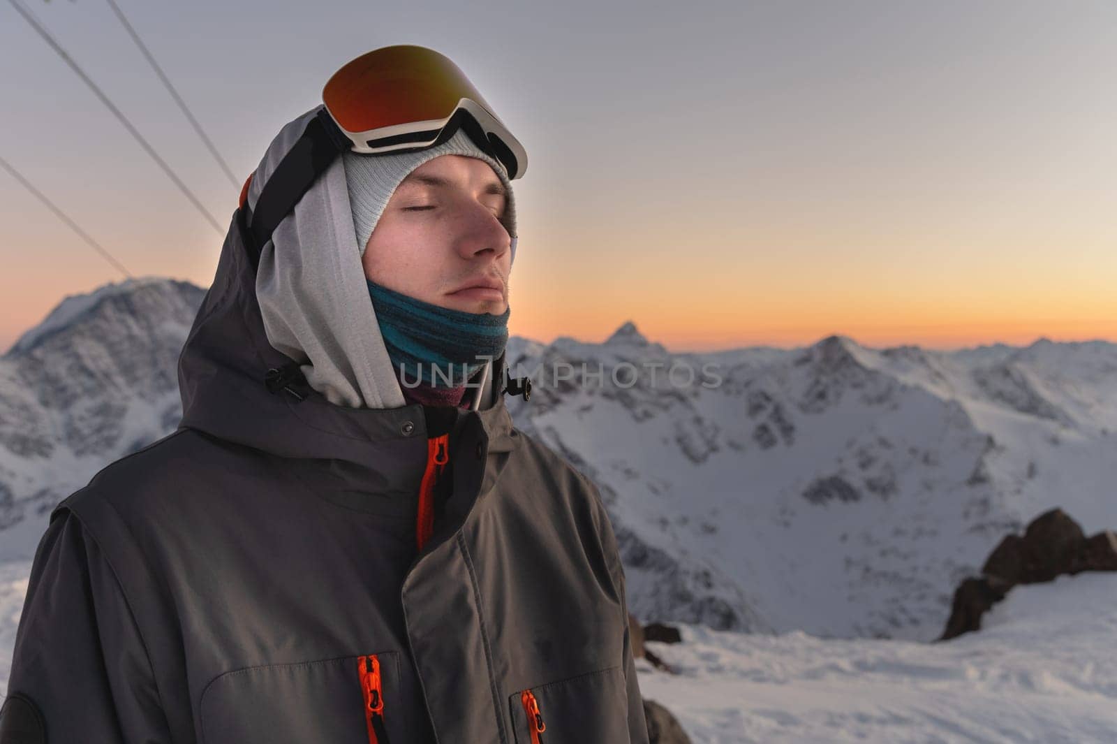 a man in ski goggles against the backdrop of snow-capped mountains and blue sky during sunset. eyes closed, enjoyment and happiness from silence and outdoor recreation.