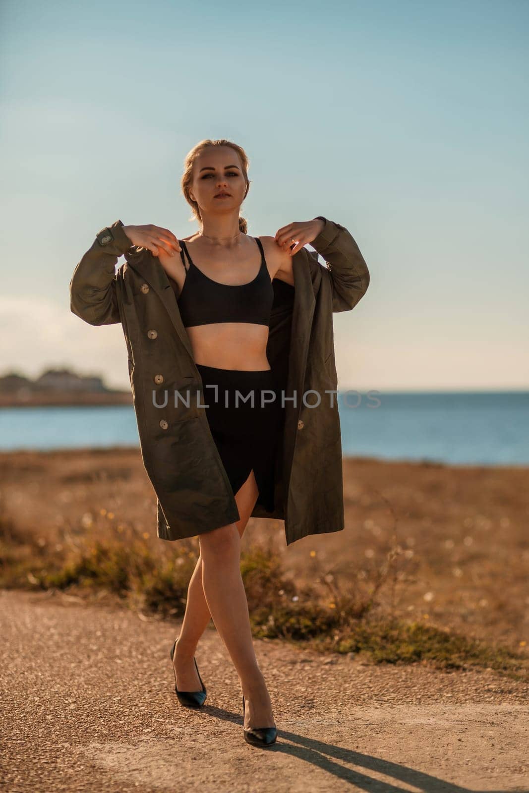 Portrait blonde sea cape. A calm young blonde in an unbuttoned khaki raincoat walks along the seashore, under a raincoat a black skirt and top.