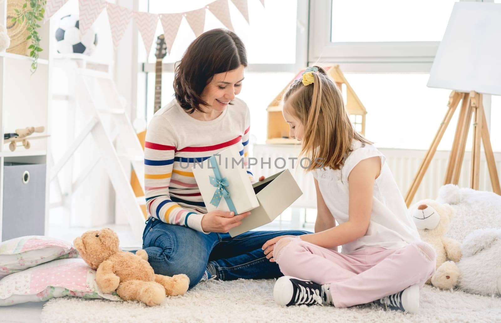 Pretty woman opening box with present from cute little girl in playroom