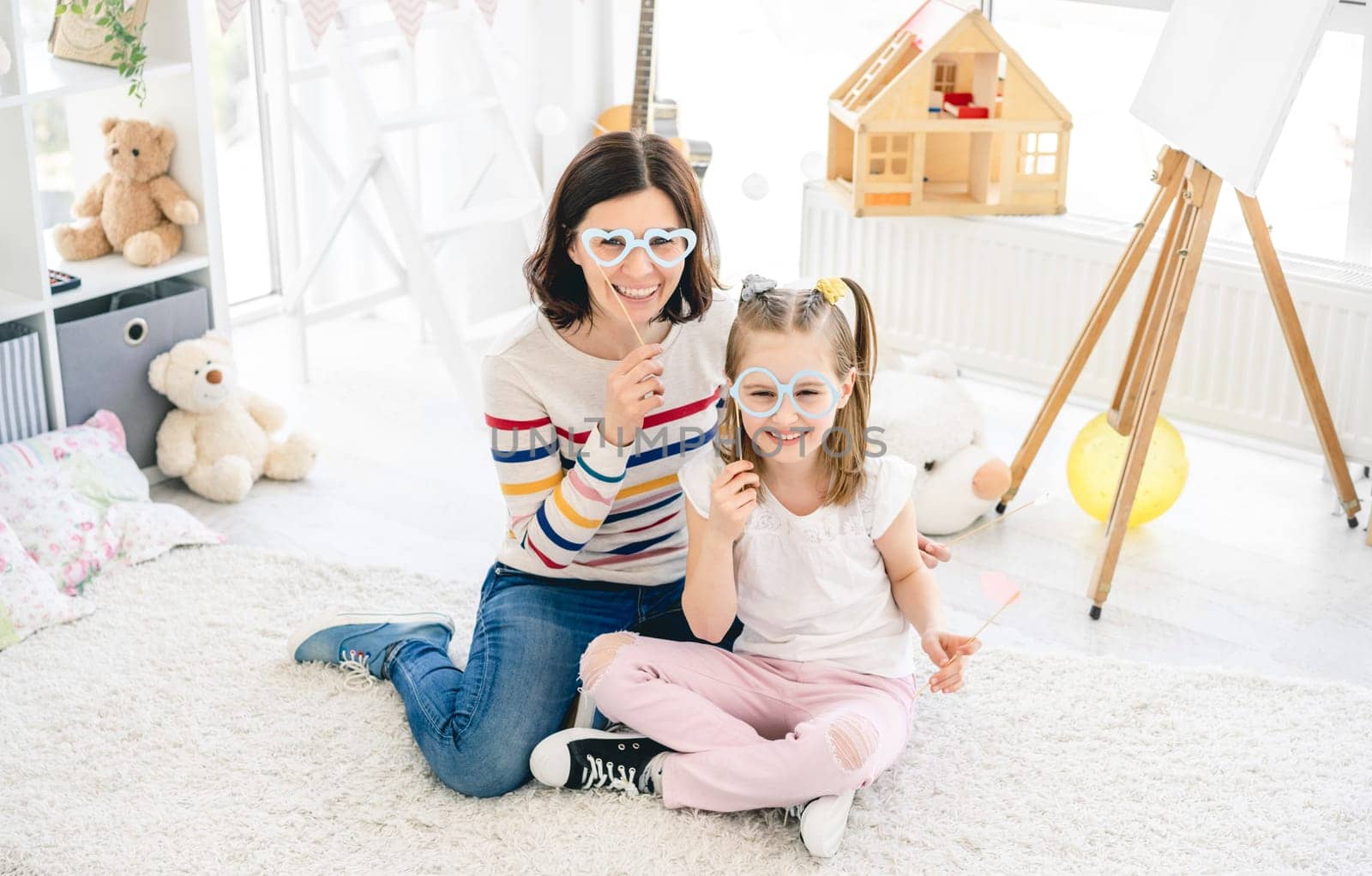 Cute little girl and mother holding stick glasses in kids room