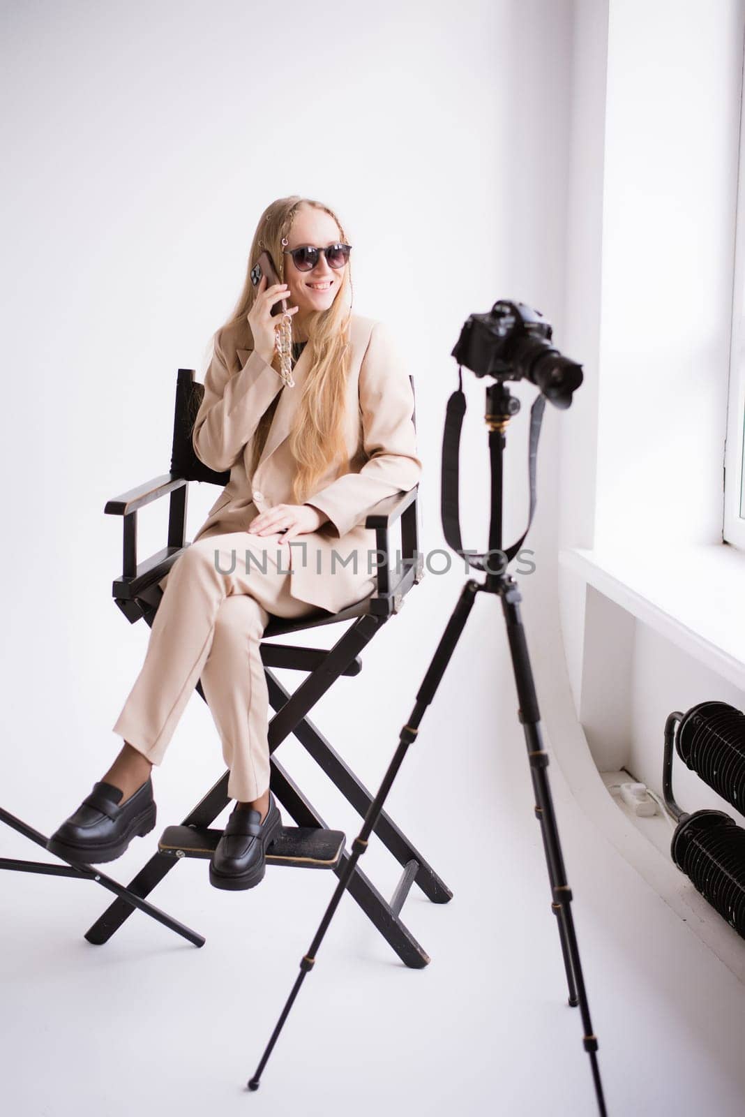 A woman producer videographer, a blonde assistant with a camera and smartphone, happy talking on the phone and smiling in the studio. Wearing a formal nude pantsuit on a white isolated background.