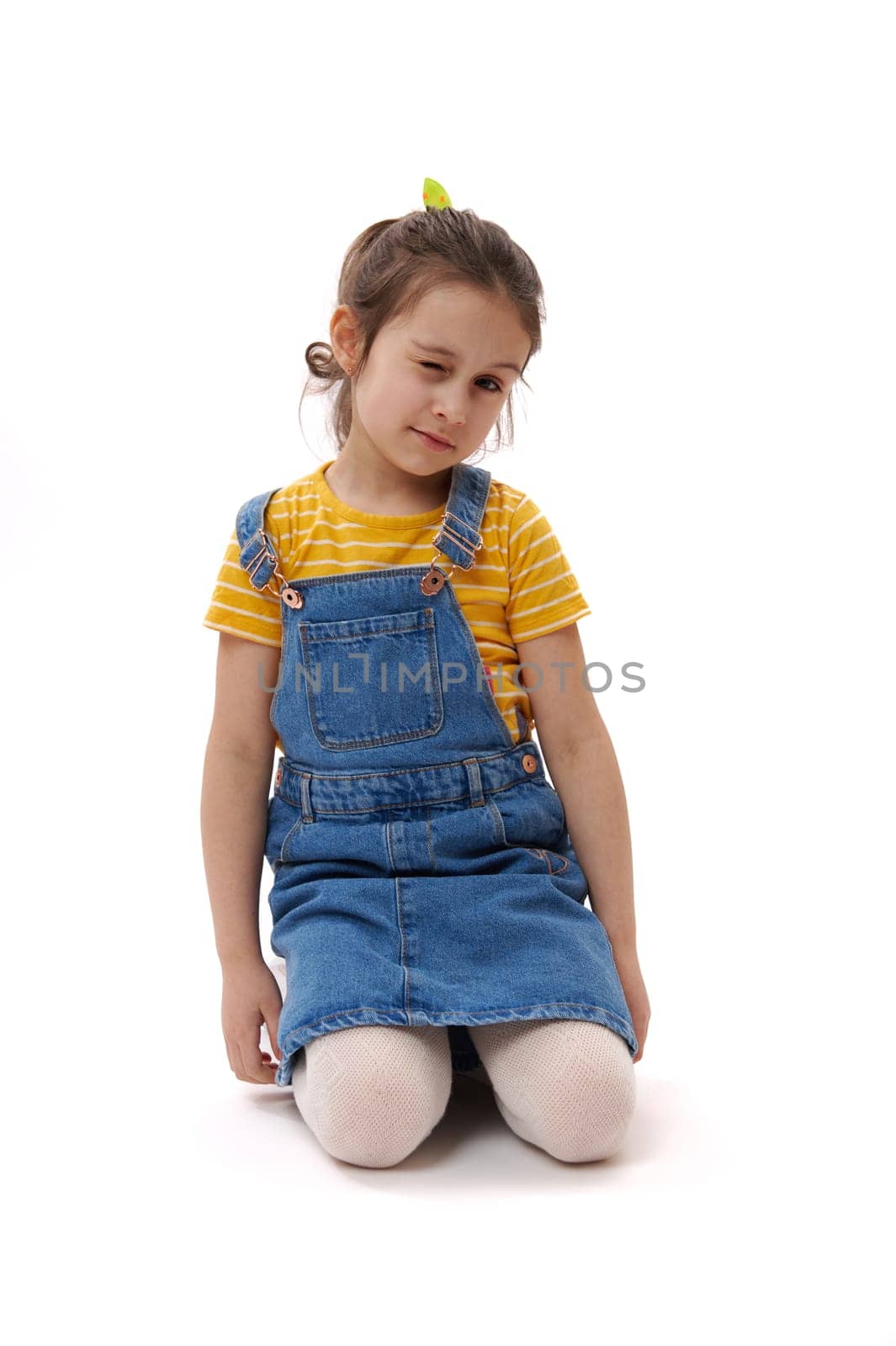 Caucasian lovely little child girl, wearing yellow t-shirt and blue denim overalls, making faces, blinking looking at camera, sitting on her knees on isolated white background. Vertical studio shot