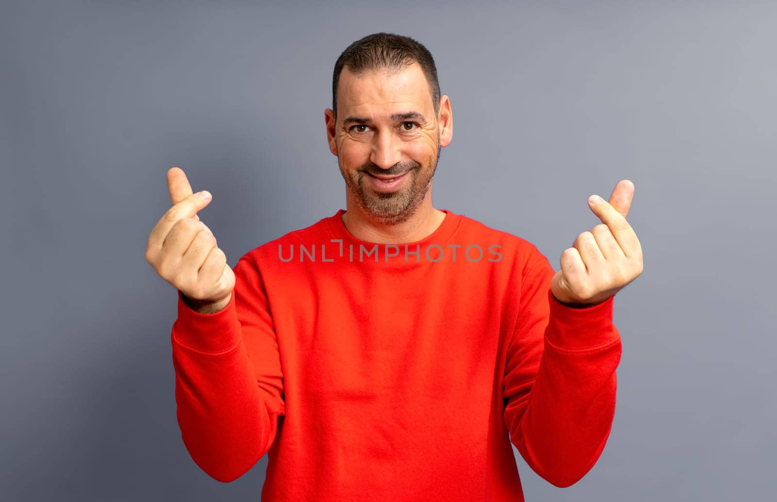 Bearded hispanic man in his 40s wearing a red sweater making the money gesture rubbing his fingers with a greedy face, isolated over gray background