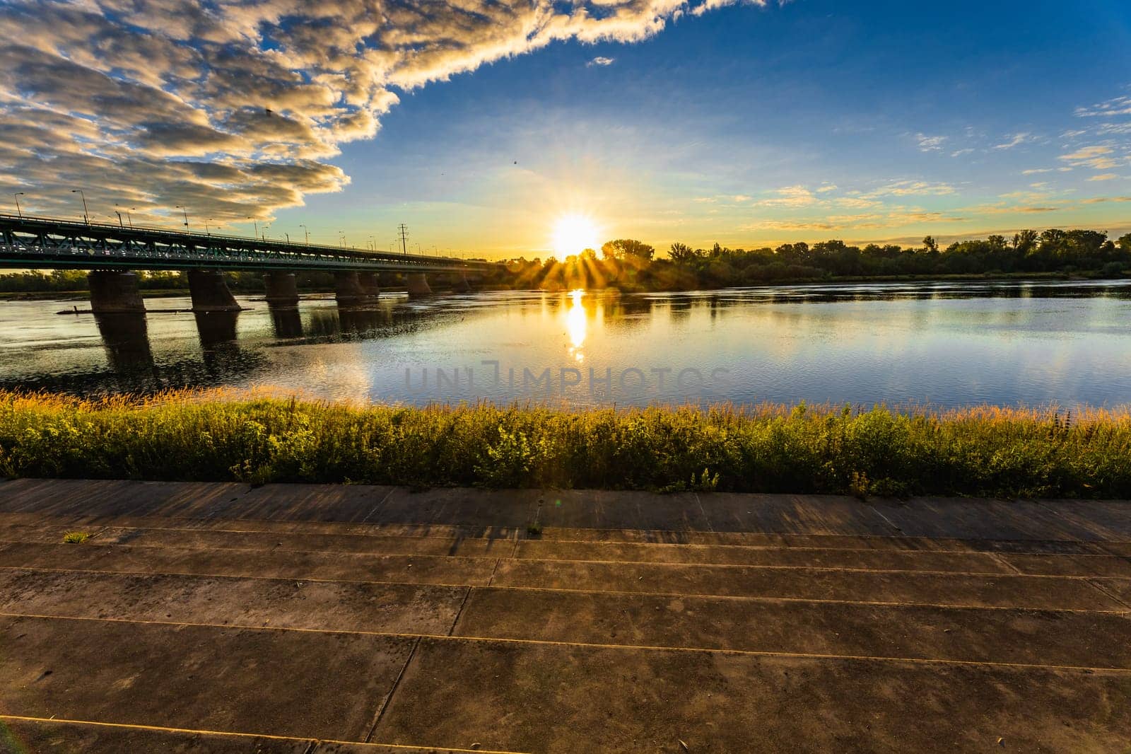 A beautiful sunrise over a wide river next to a long green bridge seen from the steps of the city promenade