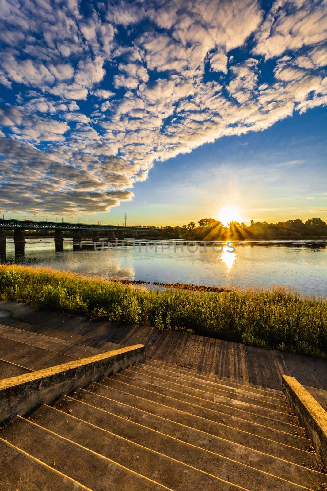 A beautiful sunrise over a wide river next to a long green bridge seen from the steps of the city promenade by Wierzchu