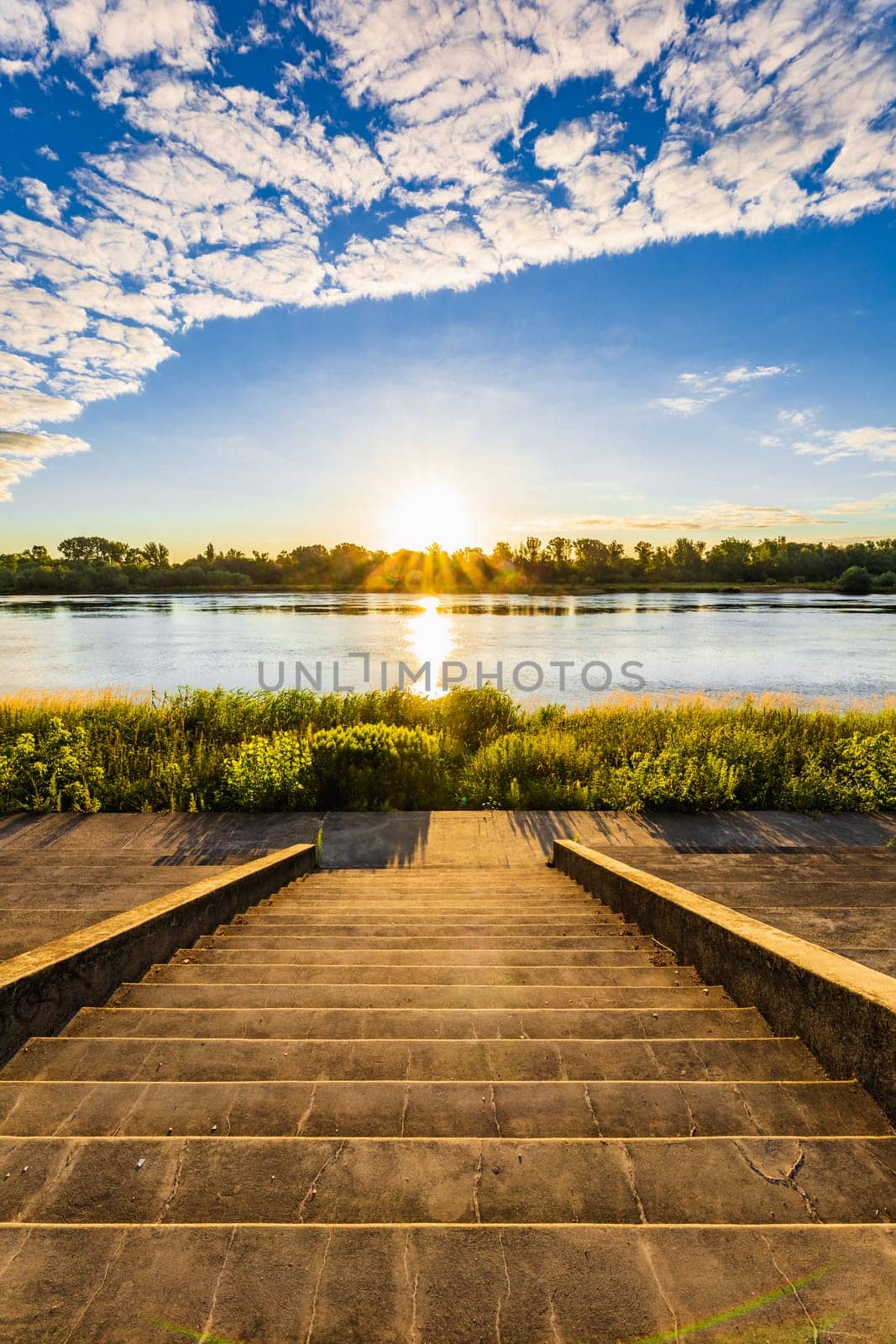 A beautiful sunrise over a wide river next to a long green bridge seen from the steps of the city promenade