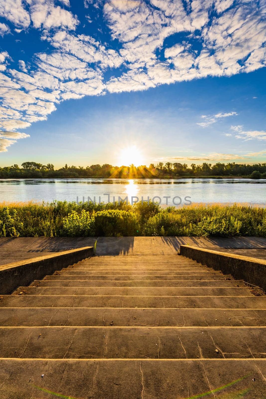 A beautiful sunrise over a wide river next to a long green bridge seen from the steps of the city promenade