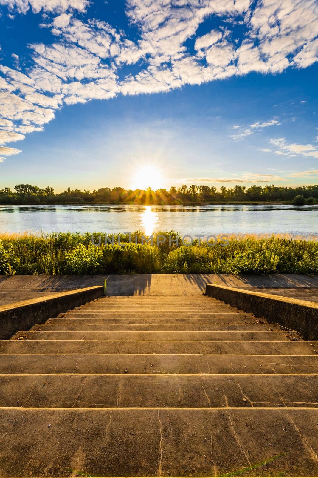 A beautiful sunrise over a wide river next to a long green bridge seen from the steps of the city promenade by Wierzchu