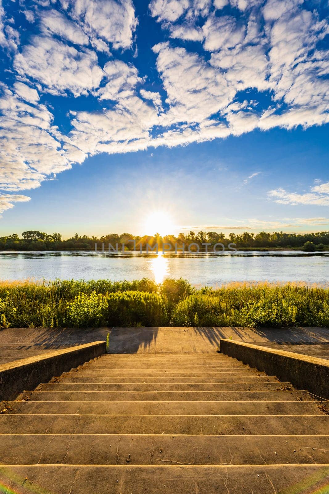 A beautiful sunrise over a wide river next to a long green bridge seen from the steps of the city promenade