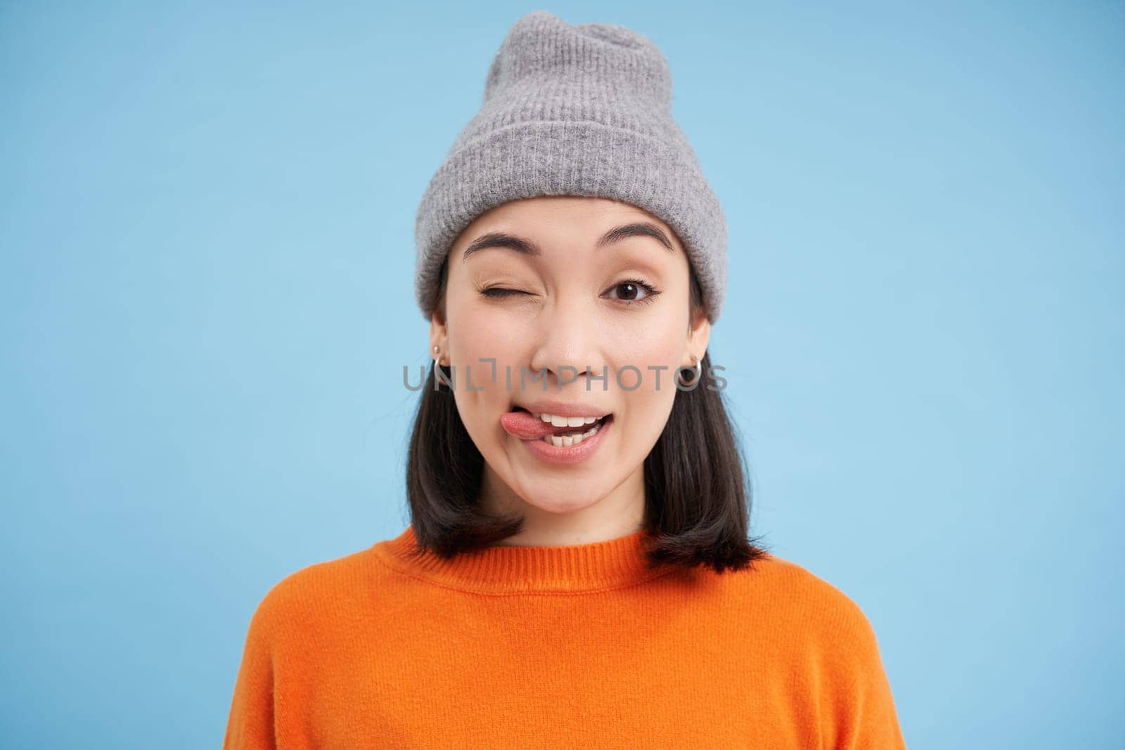 Stylish asian woman in beanie, shows tongue and winks at you, looks carefree, stands in orange sweatshirt over blue background.