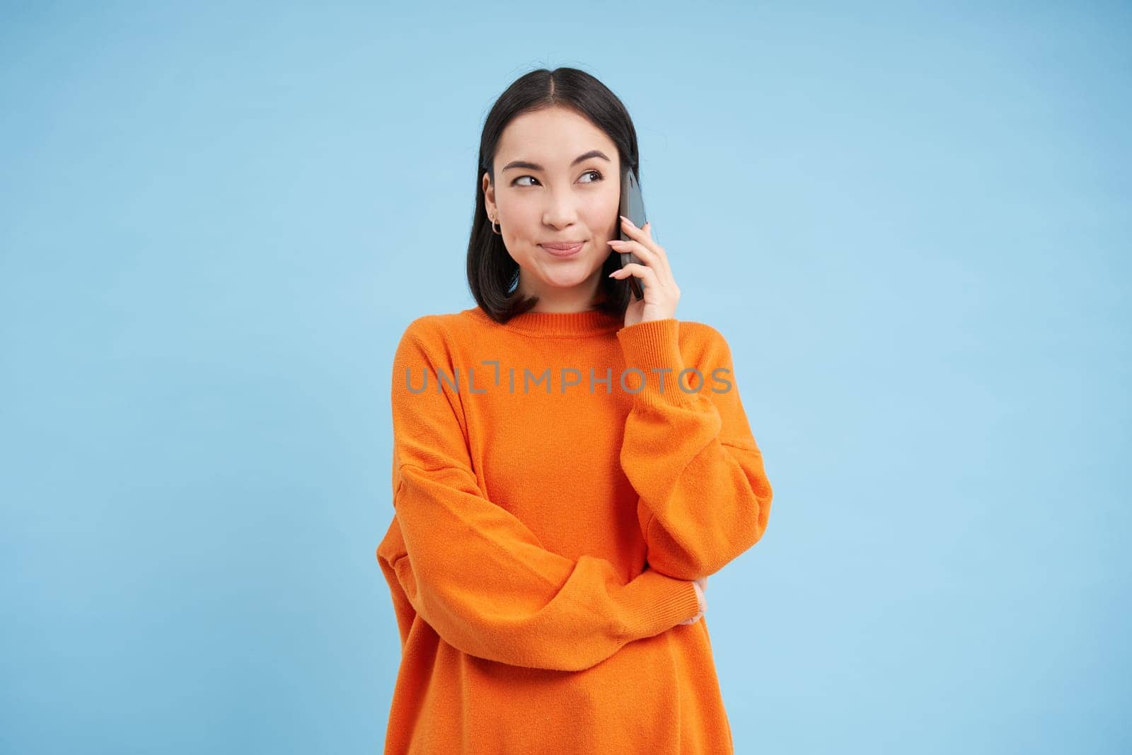 Korean girl in orange sweater talks on mobile phone, speaks on cellphone with happy smiling face, orders delivery, consults with someone on telephone by Benzoix