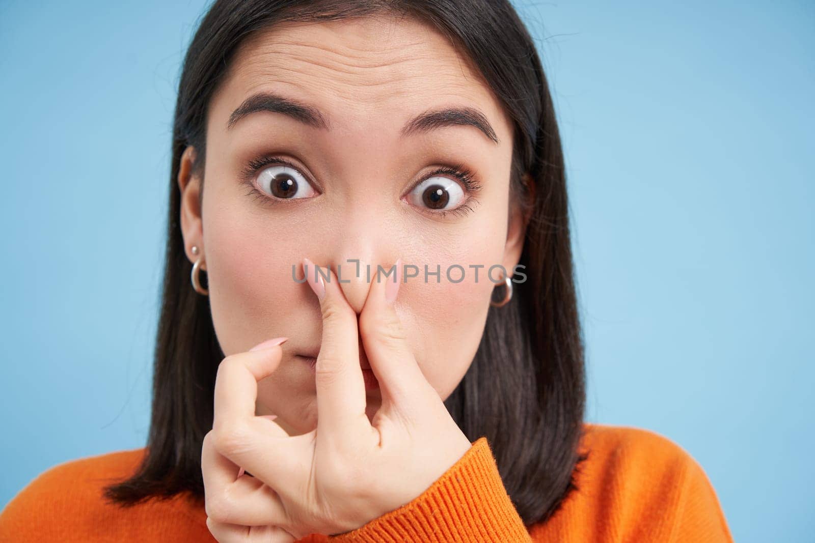 Close up portrait of japanese woman shuts her nose from bad smell, stands over blue background.