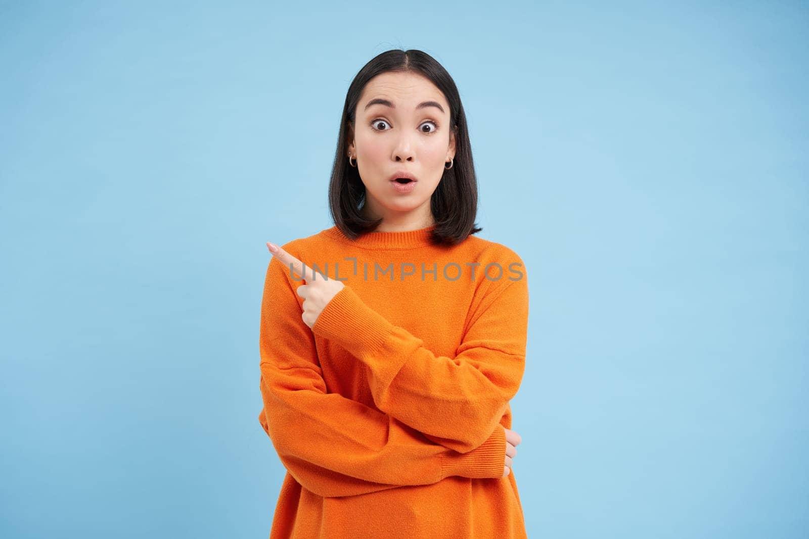 Surprised asian girl points left, shows advertisement with amazed face, shocked by promo offer aside on blue background.