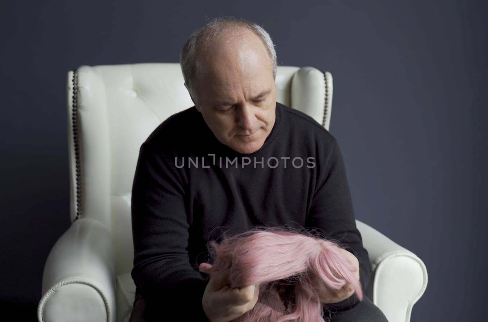 A middle-aged man sits in a chair and looks at a pink wig. Baldness problem.