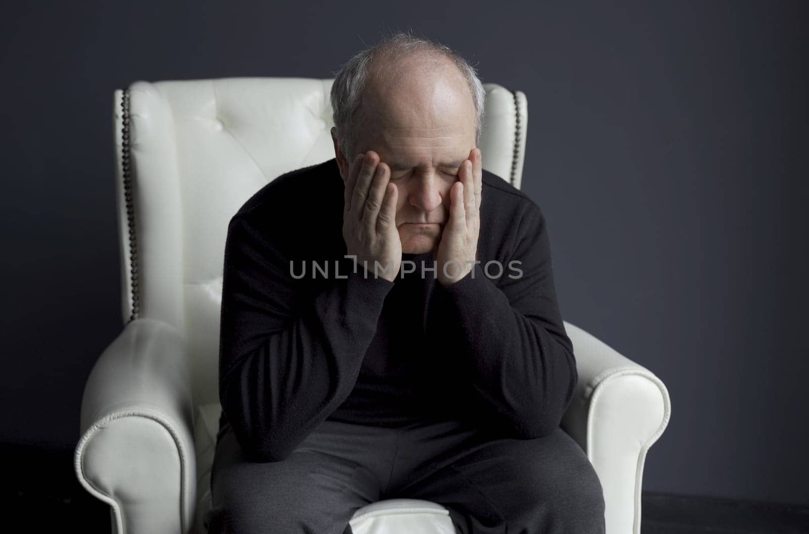 A man in a black turtleneck is sitting in a white chair, rubbing his face with his hands, holding his head. Depression, headache. Medical concept.