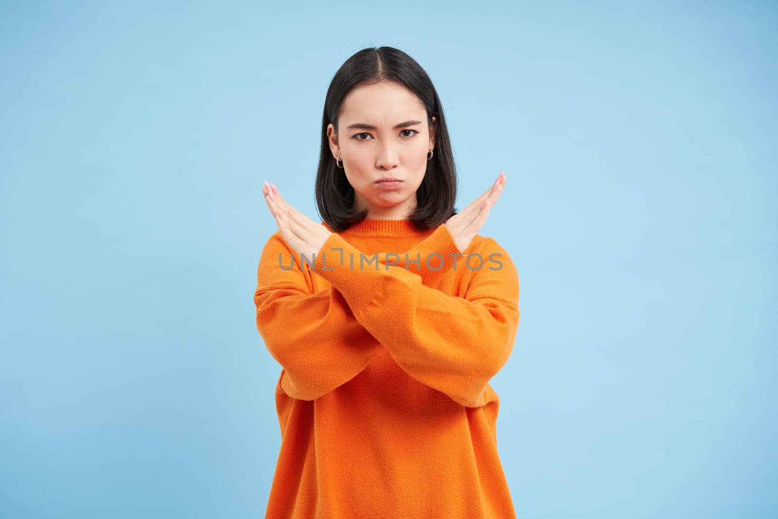 Image of serious korean woman shows cross hands gesture, stop sign, taboo, disapproves bad action, stands in orange shirt over blue background.