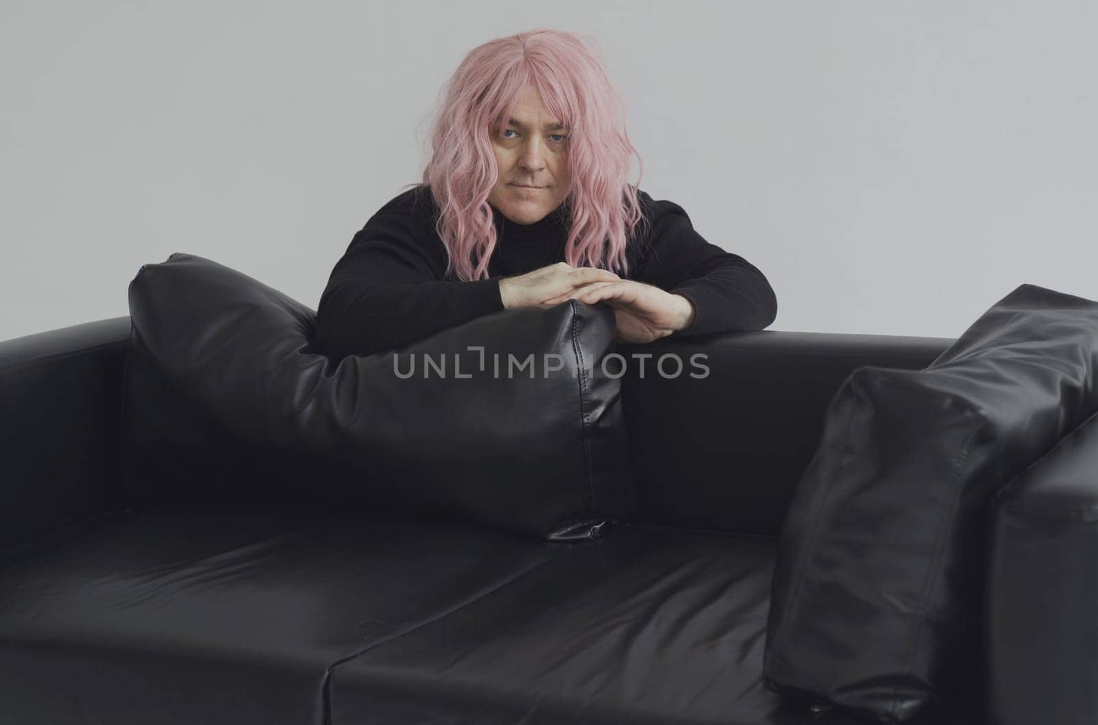 Portrait of a man in a pink wig leaned on the sofa, posing. White background.