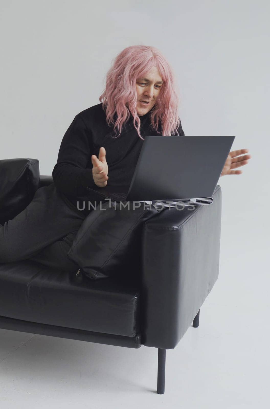 Portrait of a smiling man in a pink wig, sits on a sofa, communicates via video link on a laptop. White background. Vertical frame.