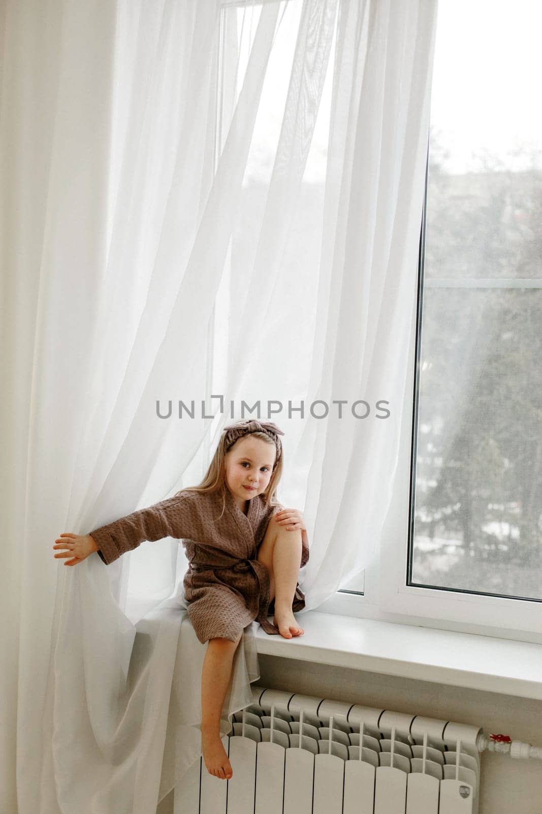 Portrait of a little girl in a brown dressing gown, who is sitting on the windowsill.