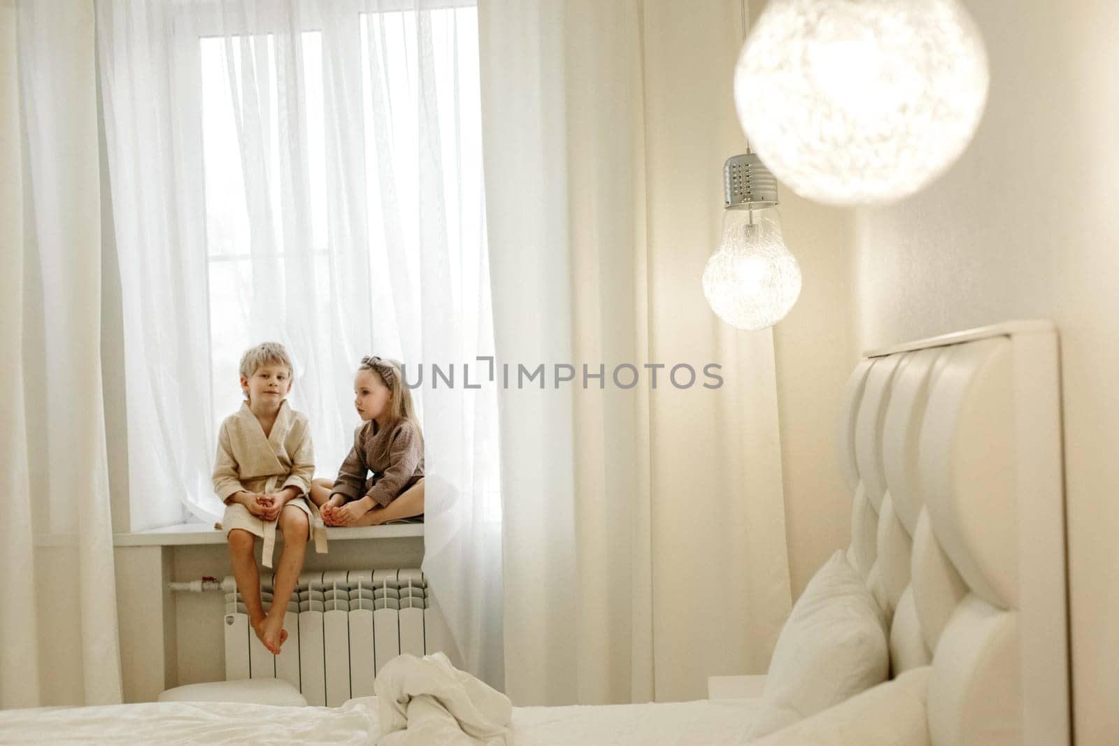 A boy and a girl are sitting on the windowsill in the bedroom.