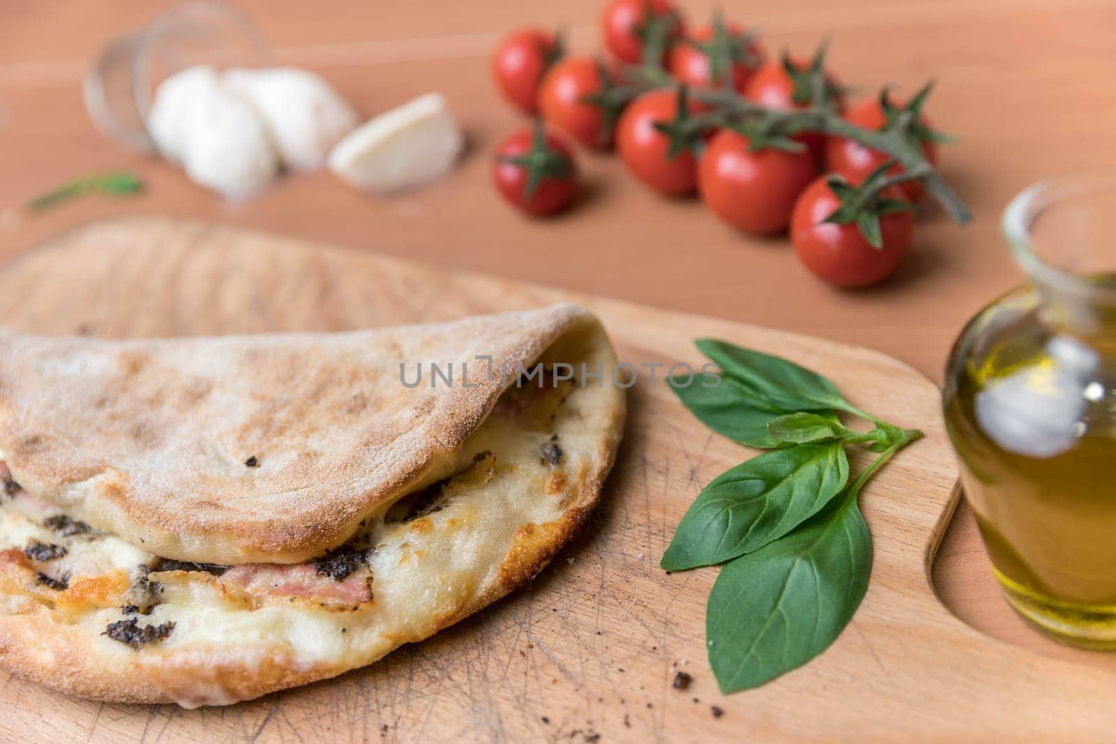 Italian food, closed calzone pizza on rustic wooden background. On background small red tomatoes, mozzarella, basil and crude oil.