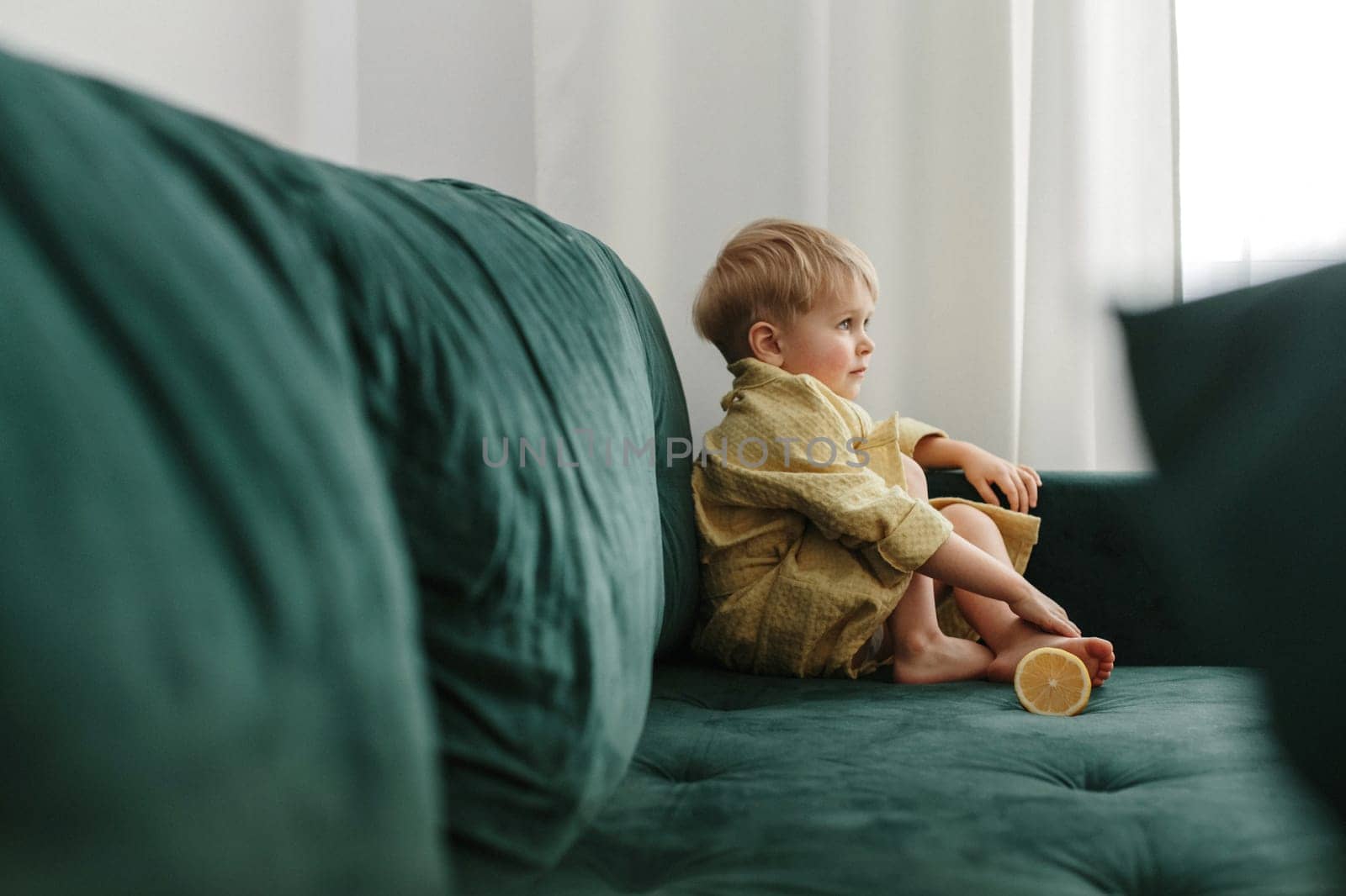 A boy in a yellow bathrobe sits on a green sofa, half an orange lies next to him.