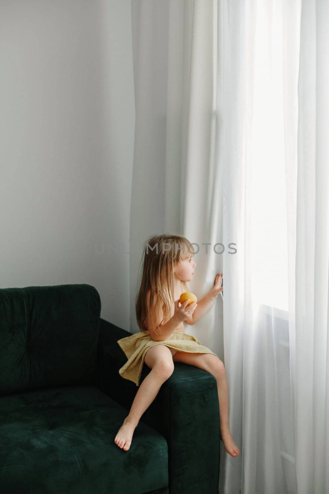 The girl sits on a green sofa, eats an orange and looks out the window.