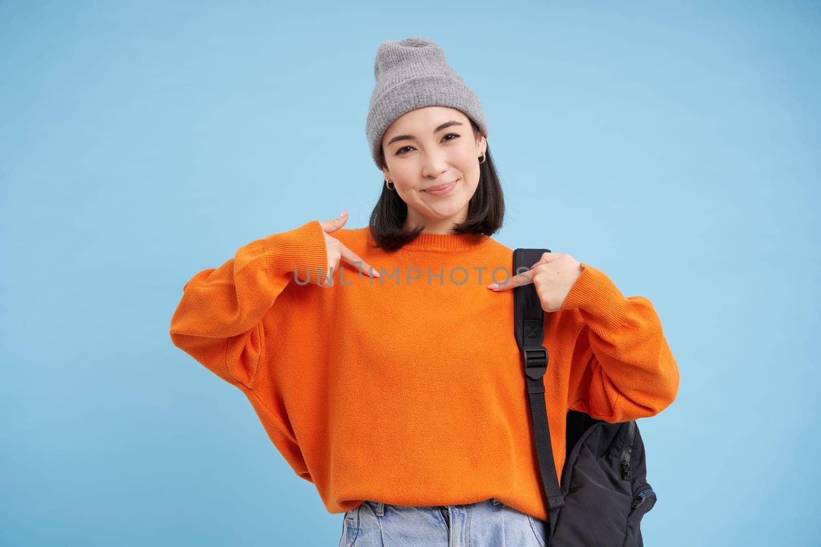 Stylish korean woman points at herself, stands in warm hat and holds backpak on shoulder, blue background. Copy space