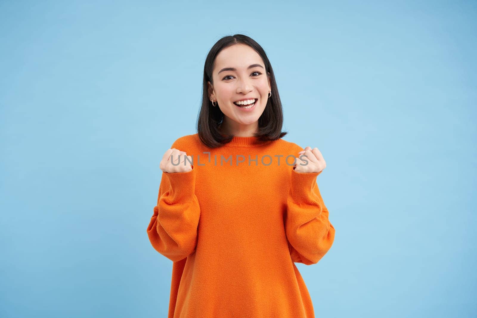 Excited young woman celebrating, jumping with hooray smiling face, winning, achieve goal, triumphing while standing over blue background by Benzoix