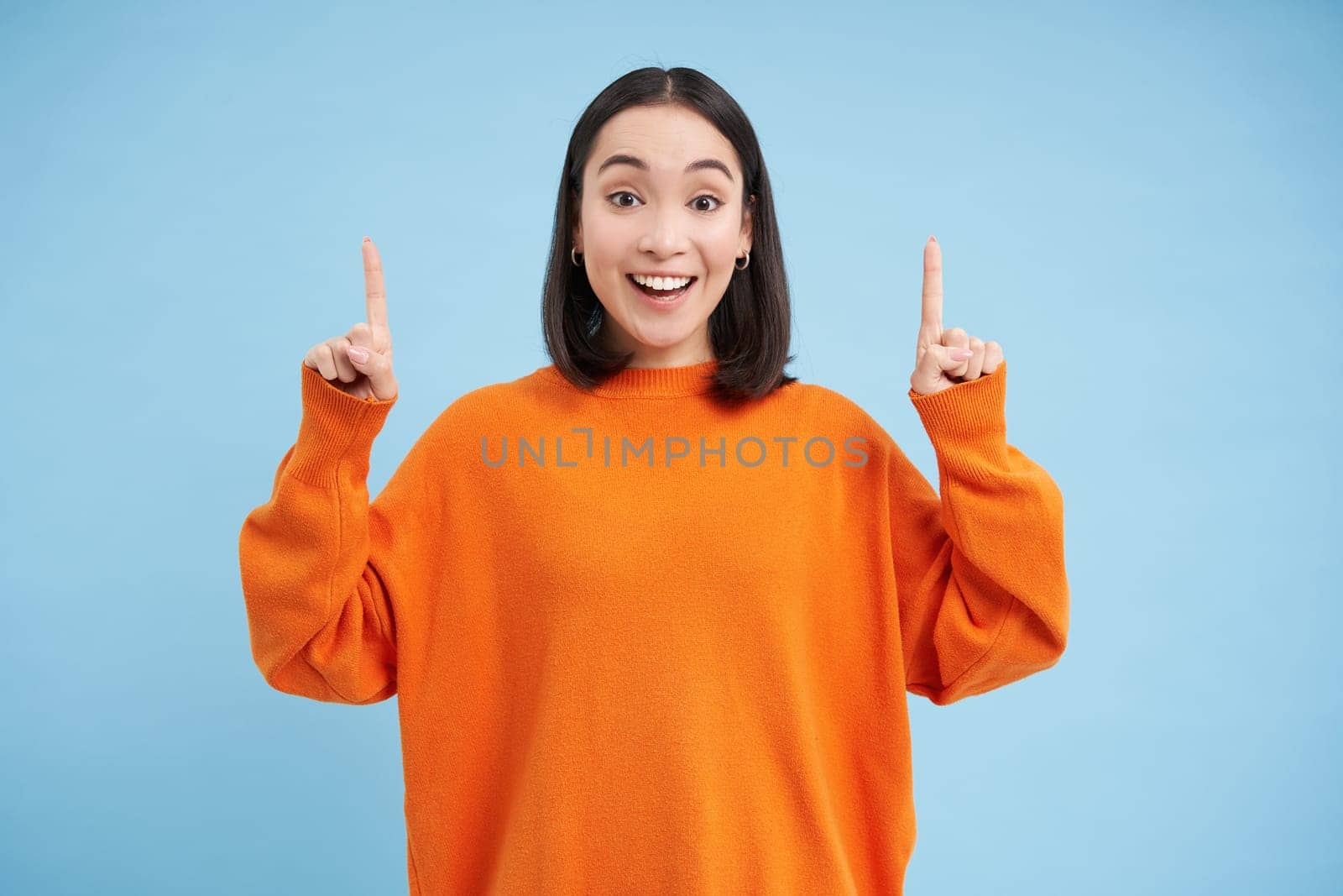 Cheerful asian woman, student shows advertisement, points fingers up and looks at promo, demonstrates promotion on top, stands over blue background.