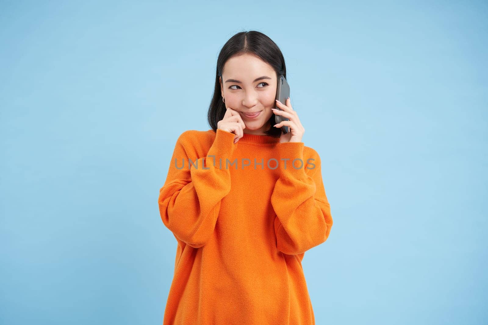 Portrait of young candid woman, asian girl talking on cellphone, holds mobile phone and speaks, stands over blue background.