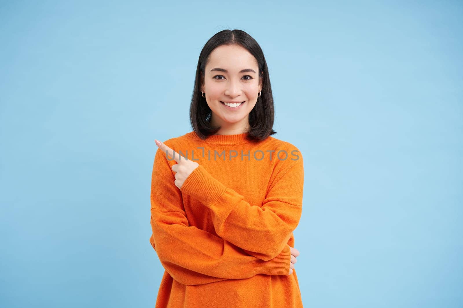 Enthusiastic asian girl, student points finger left at copy space, shows promotion offer and smiles, demonstrates advertisement, blue background by Benzoix