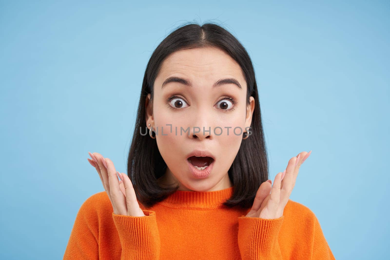 Close up portrait of amazed brunette girl says WOW, amazed by smth. Asian woman looks surpirsed at camera, impressed reaction, blue background.