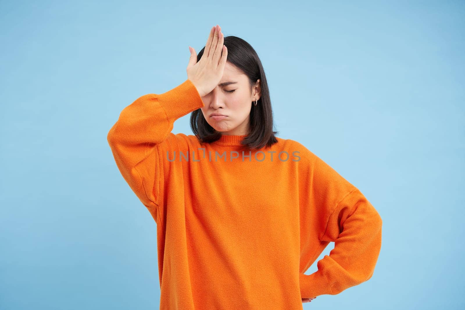 Epic fail. Disappointed asian woman slaps her forehead with upset face, forgot smth, annoyed, stands over blue background in orange sweatshirt.