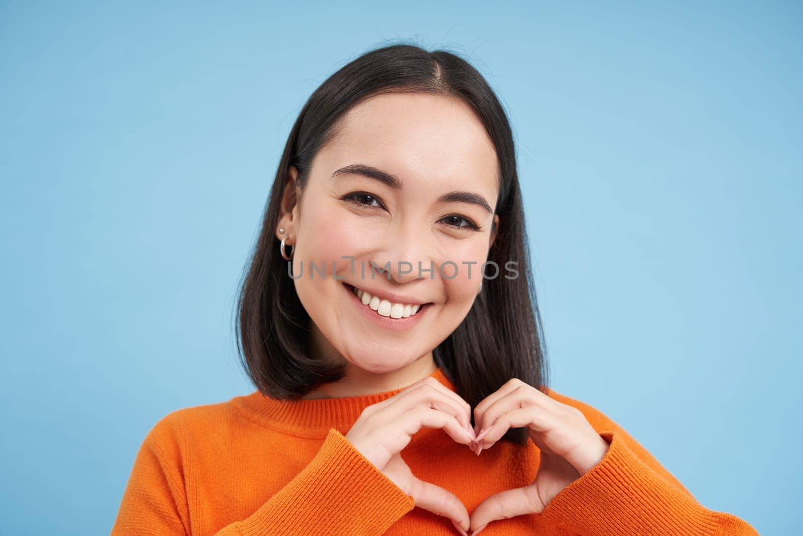 Close up portrait of lovely asian woman, shows heart sign and smiles with care and tenderness, stands over blue background.