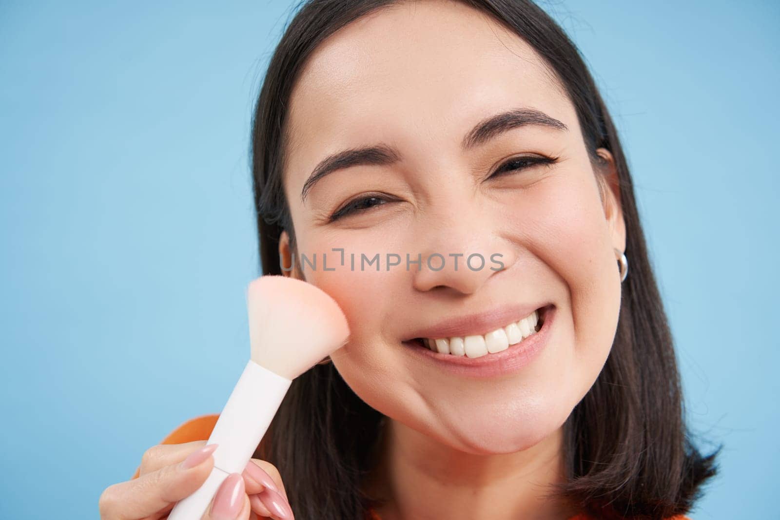 Portrait of cheerful korean girl smiles, puts on makeup with brush, stands over blue background by Benzoix
