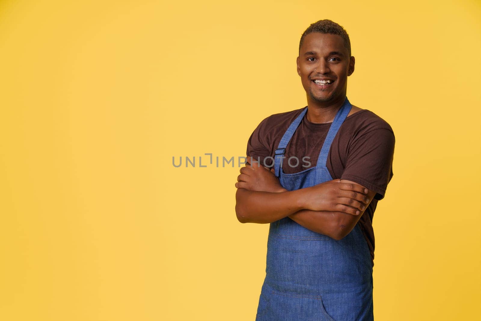Cheerful African American cook wearing blue apron smiles in front of yellow background with copy space, suggesting concept of professional cook or chef in culinary or kitchen-related profession. High quality photo