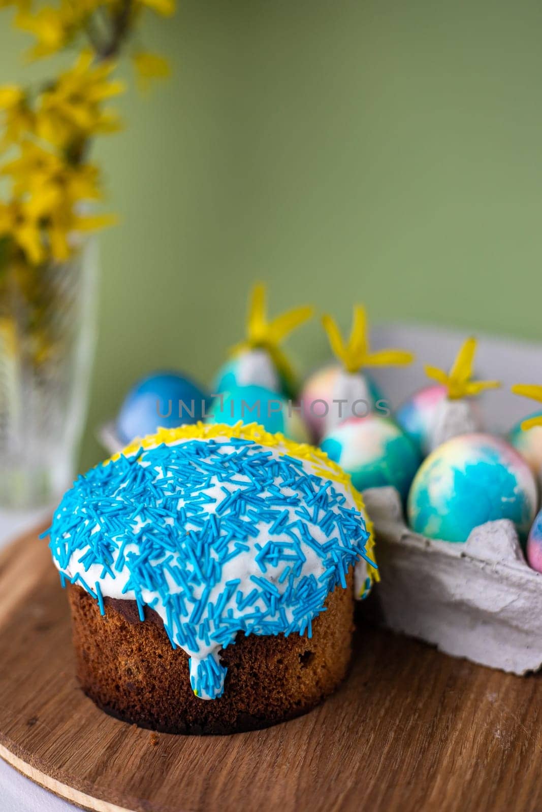 Easter composition with yellow-blue Easter cakes, sprinkles, Easter multi-colored eggs. wooden stand and spring flowers on a yellow background. copy space by Anyatachka