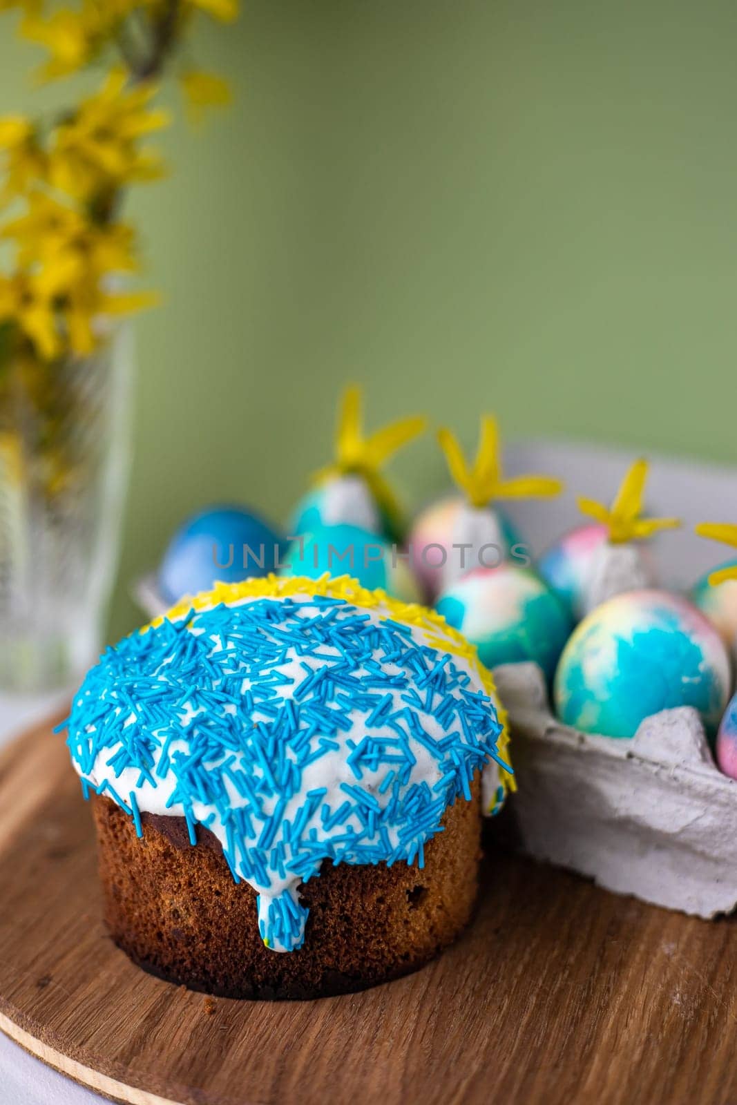 Easter composition with yellow-blue Easter cakes, sprinkles, Easter multi-colored eggs. wooden stand and spring flowers on a yellow background. copy space.