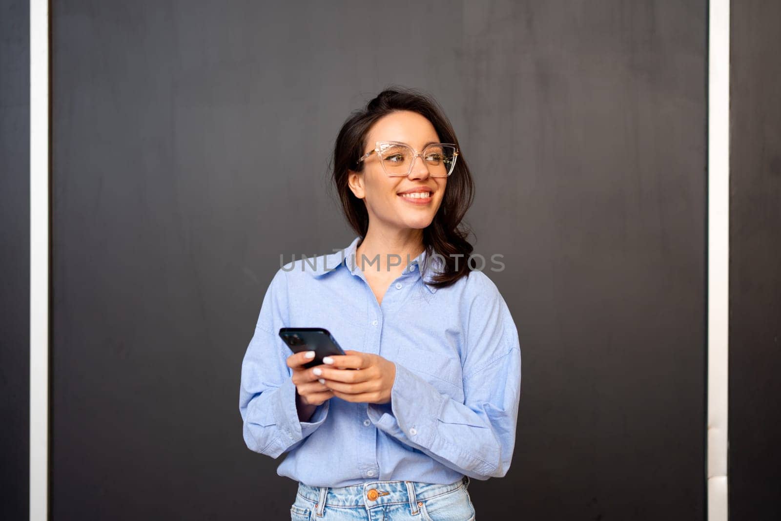 Portrait smiling woman holding smartphone in hand looking away, wear glasses, types text message on mobile phone, enjoys online communication, on black studio background. Communication concept