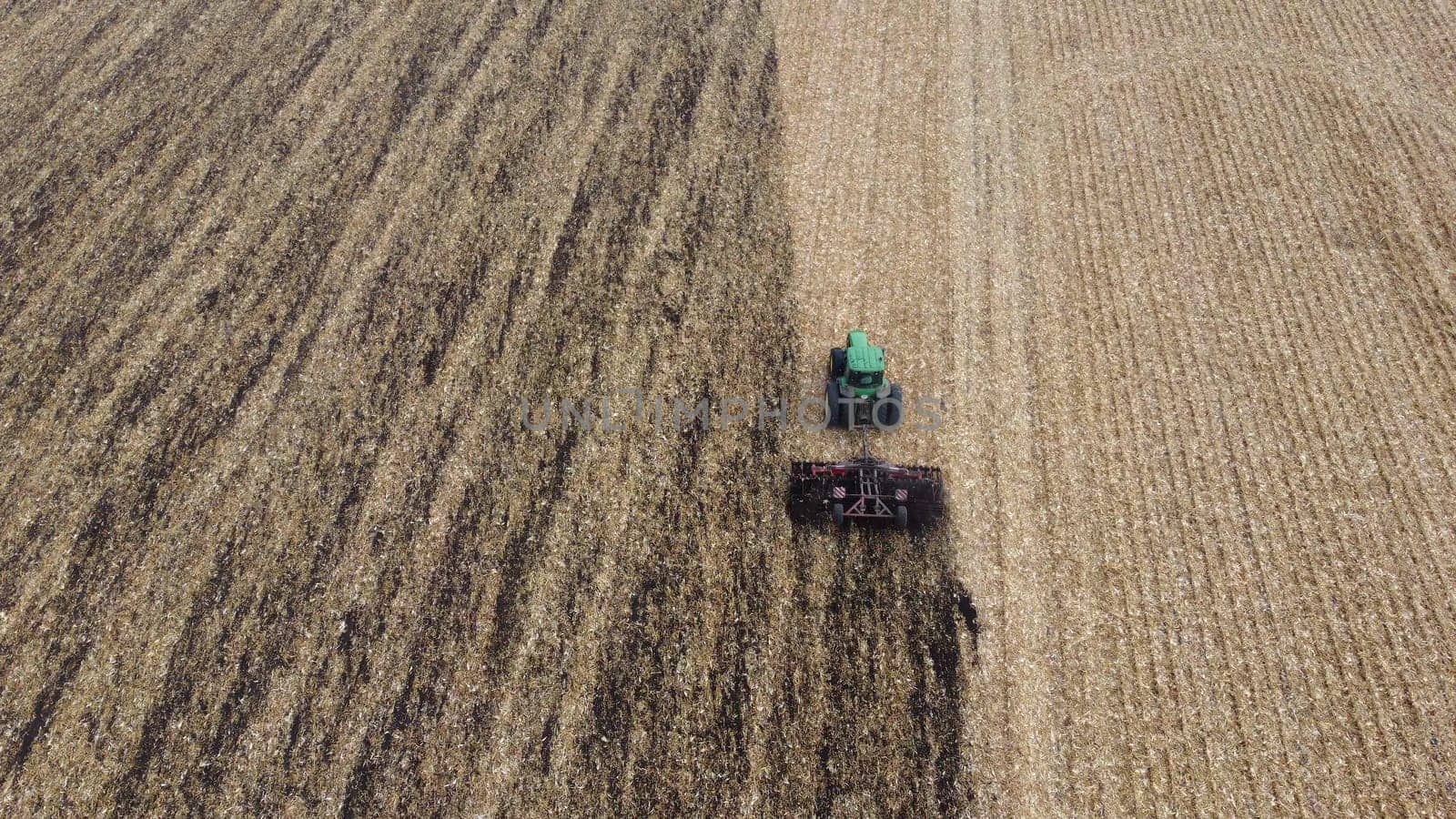 Tractor plowing the ground. Flying over green tractor that plows up ground in yellow field after harvesting wheat on autumn day. Tractor digging land. Agricultural work on field. Aerial drone view