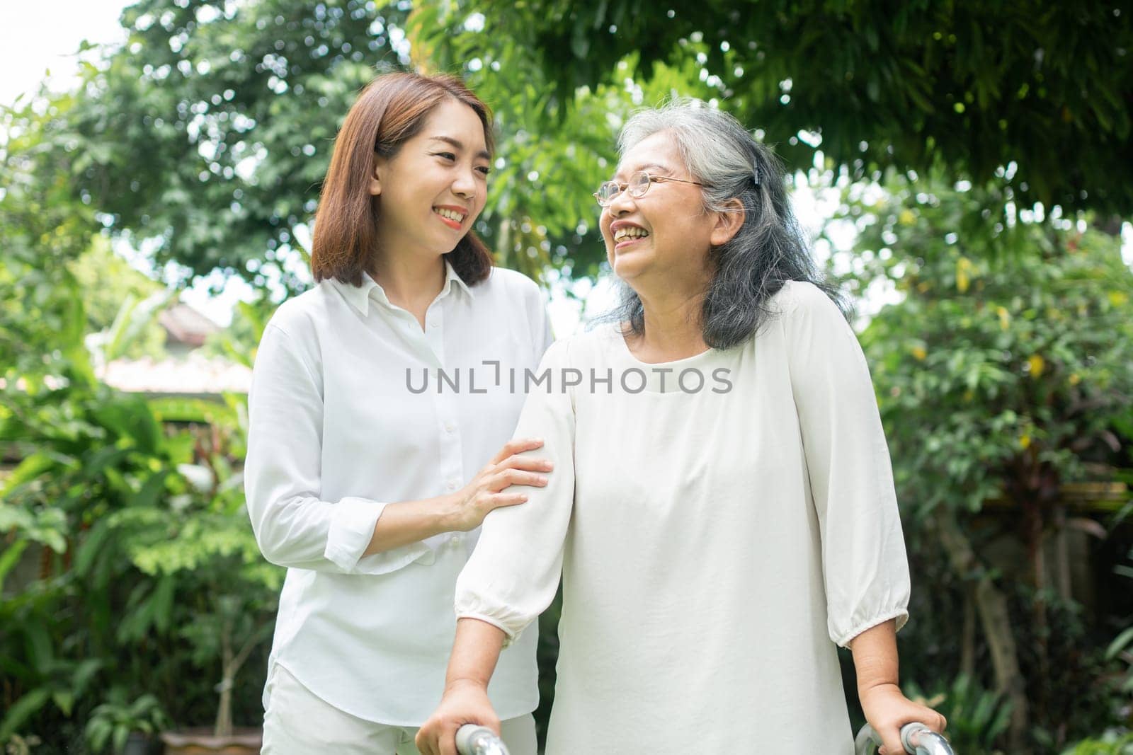 An old elderly Asian woman uses a walker and walking in the backyard with her daughter.  Concept of happy retirement With care from a caregiver and Savings and senior health insurance by PattyPhoto