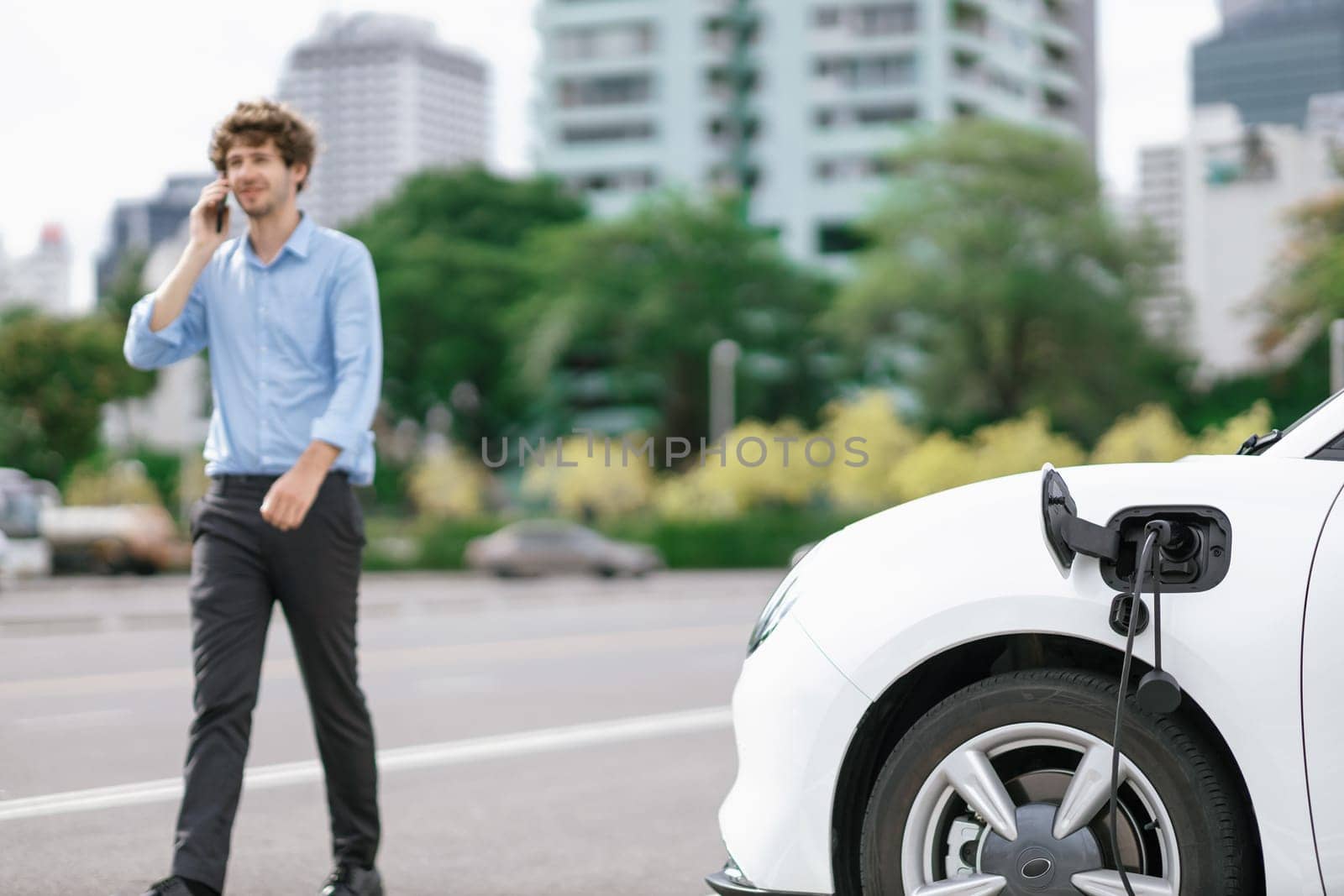 Progressive eco-friendly concept of focus parking EV car at public electric-powered charging station in city with blur background of businessman talking on the phone while recharging electric vehicle.