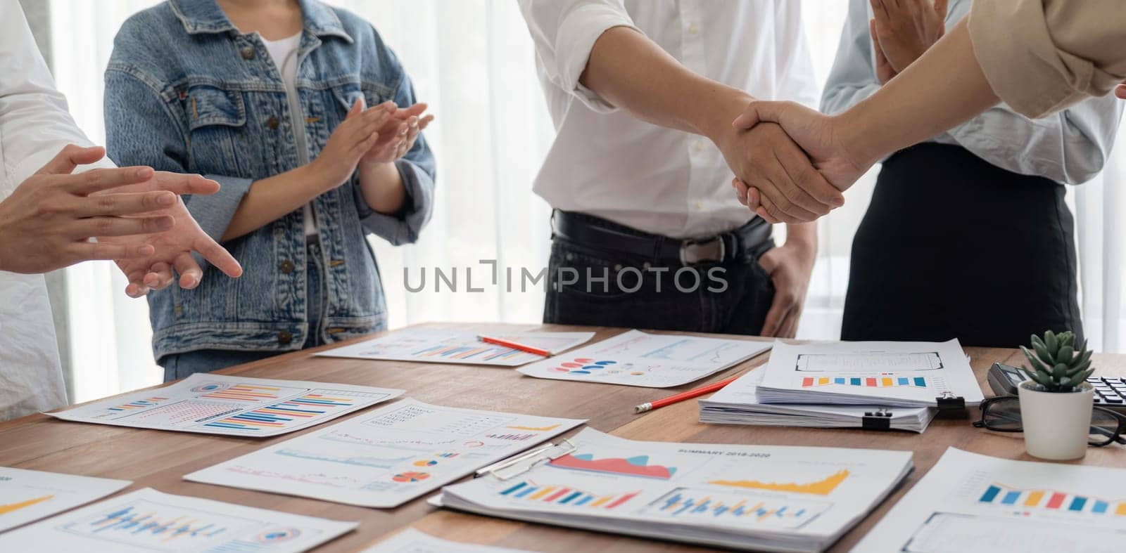 Analyst team celebrates successful data analysis with handshake. Panorama shot handshaking after adjusting attitude or brainstorm for future success in meeting room with BI dashboard paper. Scrutinize