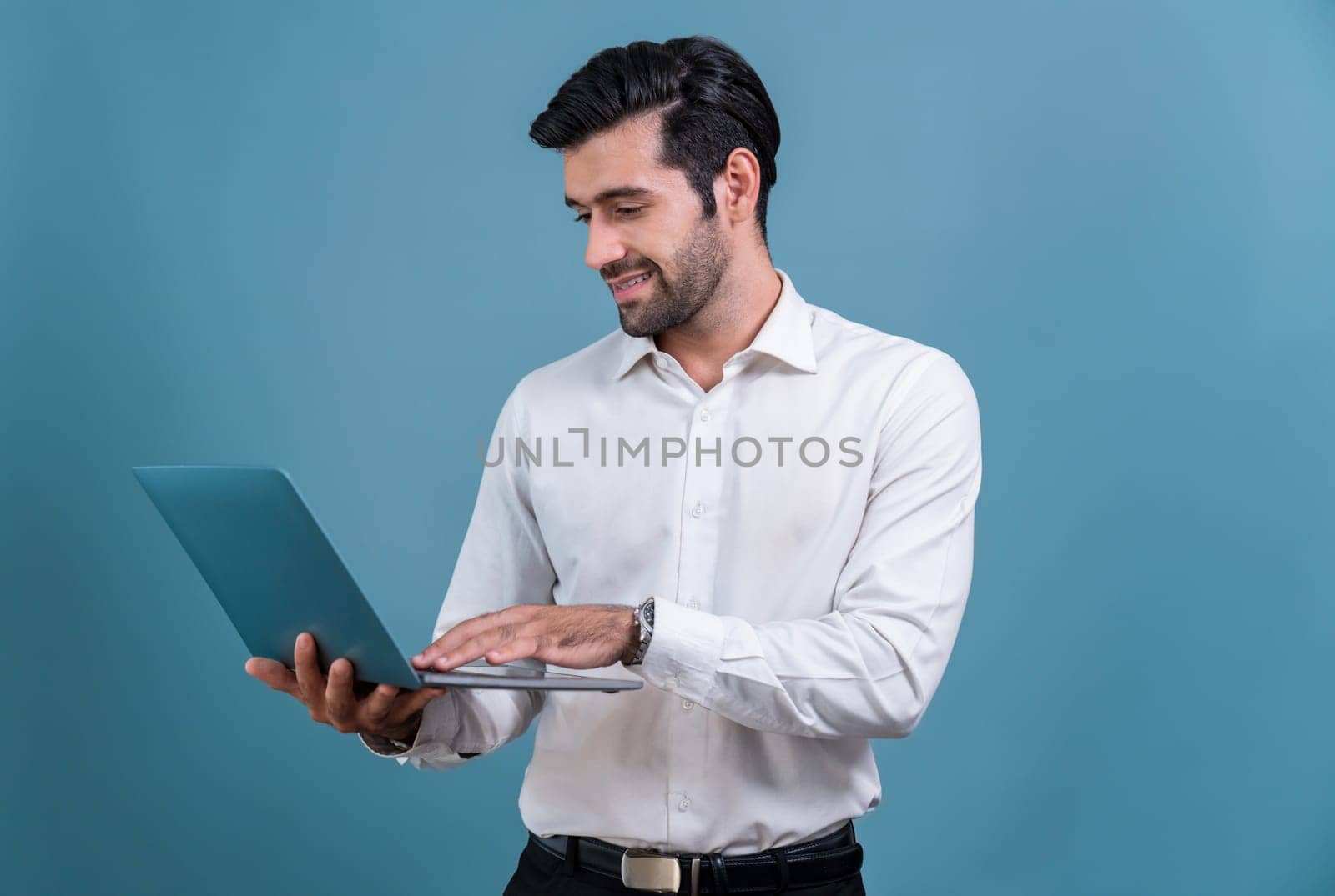 Successful businessman in black suit with innovative tech concept, standing pose and holding laptop and smiling with excitement on copyspace background for promotion or advertisement. Fervent