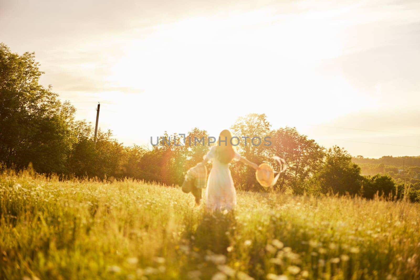 a woman in a light dress stands in a field in the warm rays of the setting sun holding a hat over her head. High quality photo