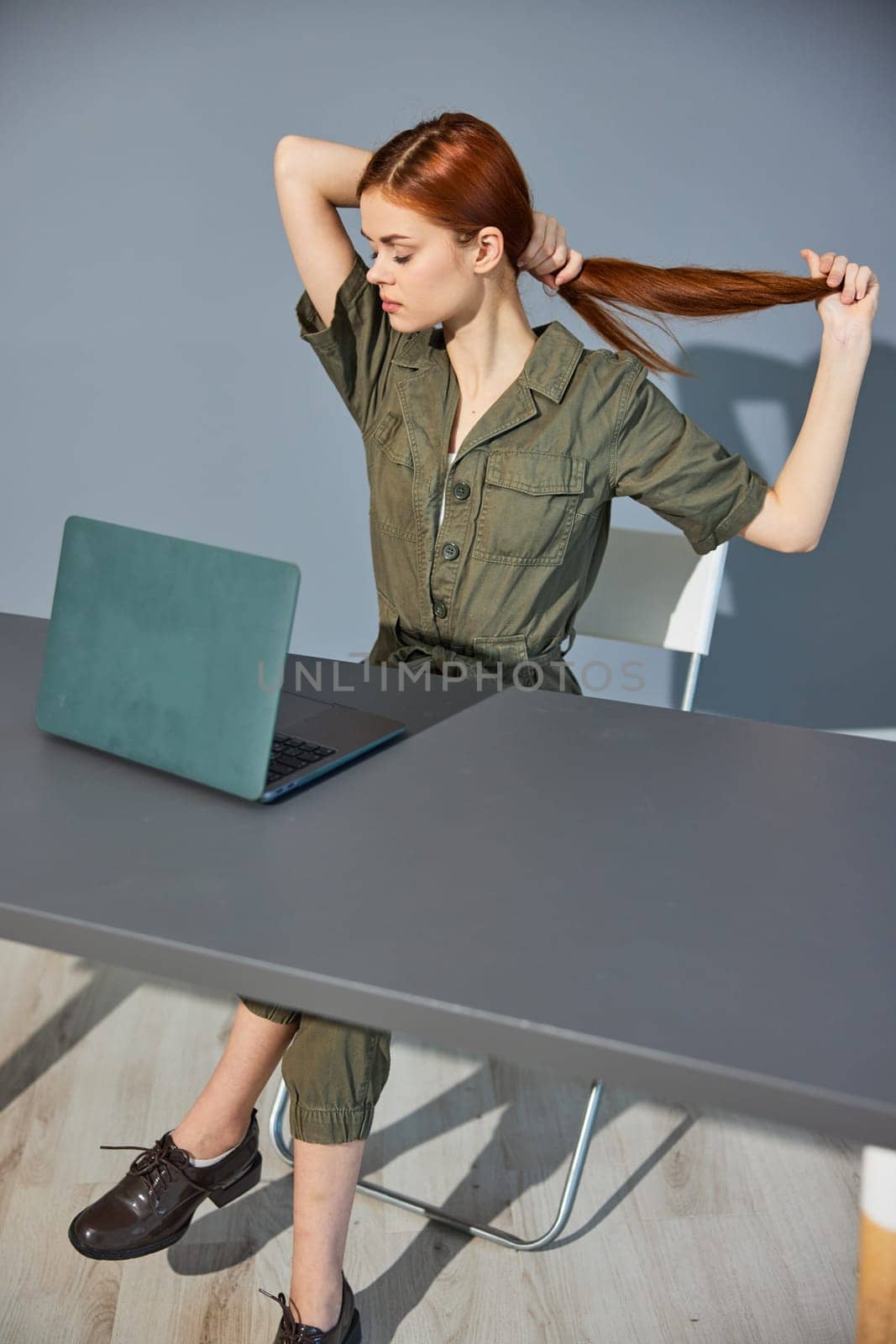 a woman in overalls sits at a laptop in the office and collects her hair with her hands. High quality photo