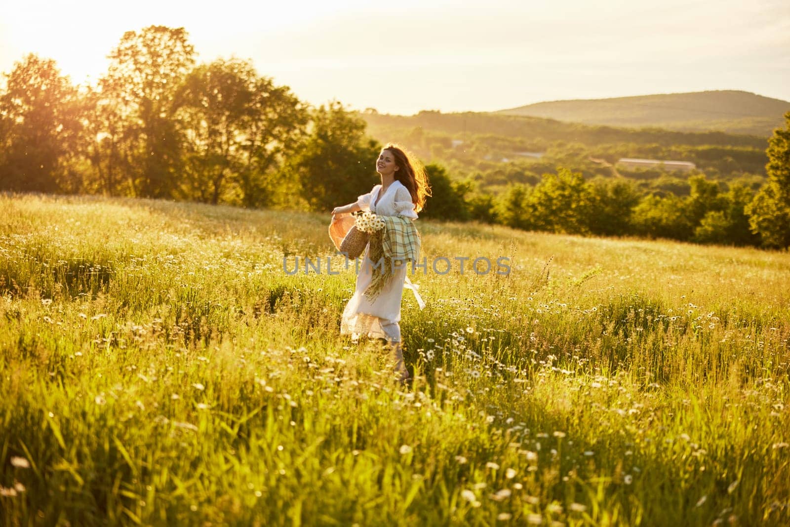 a woman in a white dress with a basket of daisies and a straw hat stands in a field during sunset in a light breeze. High quality photo