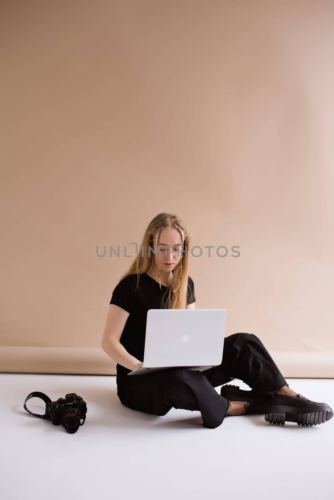 A business woman freelancer in a black is working, typing on a laptop apple and sitting on a white floor. blonde assistant in video production with computer MacBook and camera .